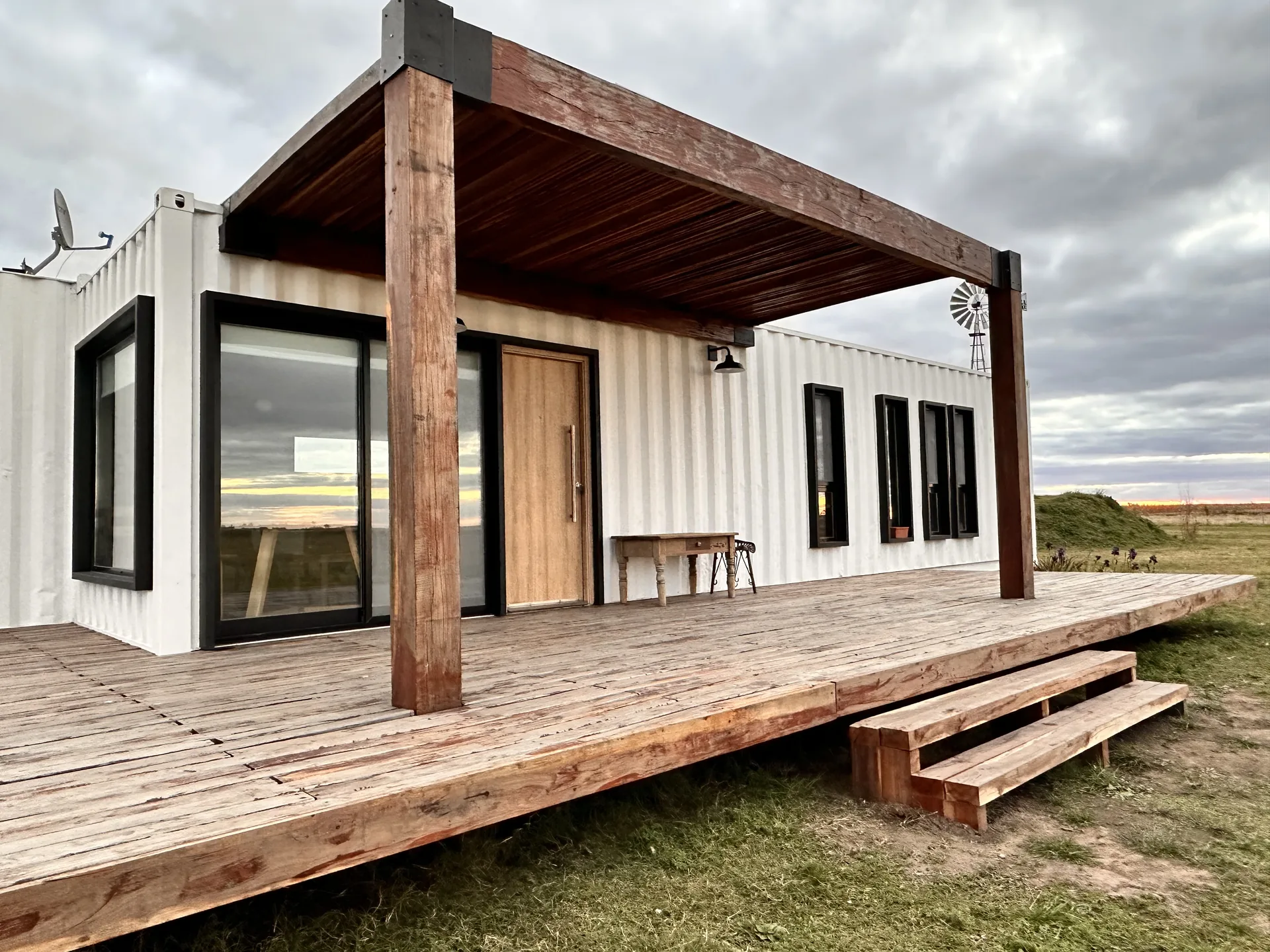 POLO Crucero: containers blancos en L con deck de madera amplio, pérgola de tirantes macizos y ventanales corredizos, con molino de campo al fondo.
